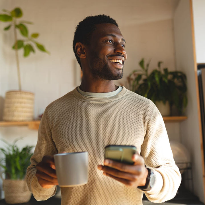 Smiling person holding a mug and phone in a cozy sunlit room with plants