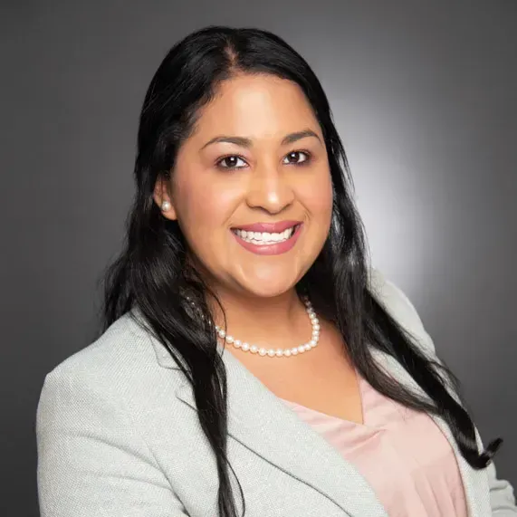 Portrait of a smiling woman in a light gray blazer and pearl necklace against a gray backdrop