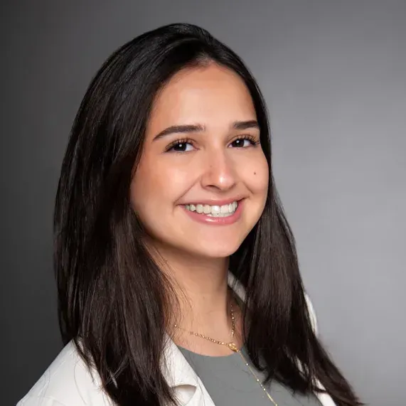 Smiling person with long dark hair in a white coat against a gray background