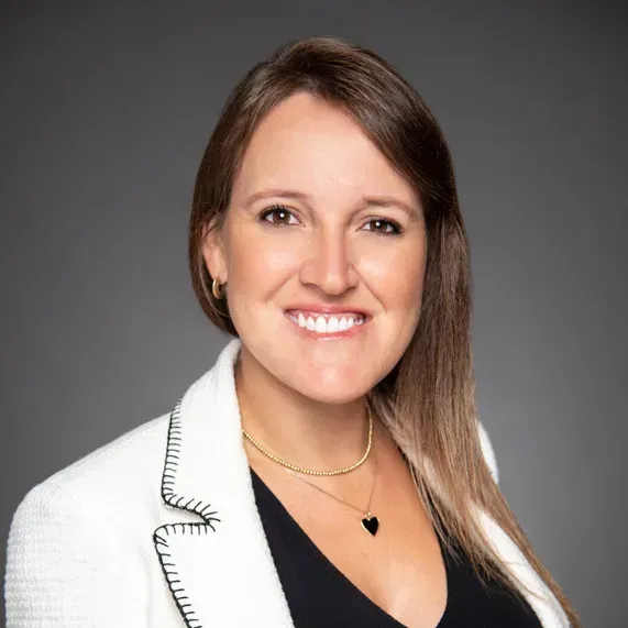 Studio headshot of a smiling woman in a white blazer against a gray background