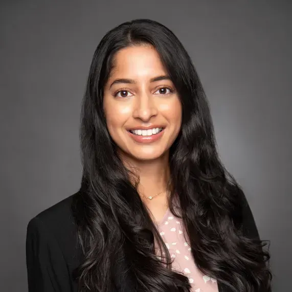 Smiling woman with long dark hair against a gray background, wearing a black blazer and pink polka-dot blouse