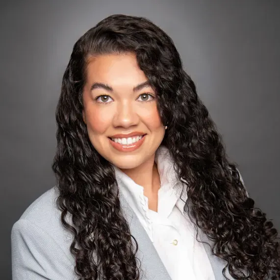 Smiling person with long curly dark hair in a white shirt against a gray background
