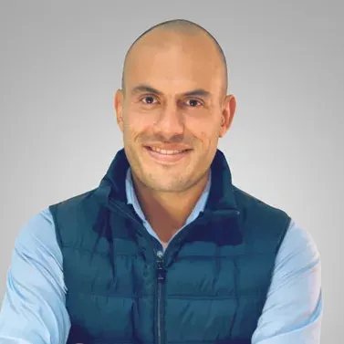 Smiling bald man in a navy vest and light blue shirt against a gray background