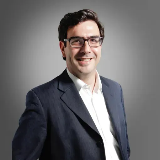 Portrait of a smiling man in glasses wearing a dark blazer and white shirt against a gray background