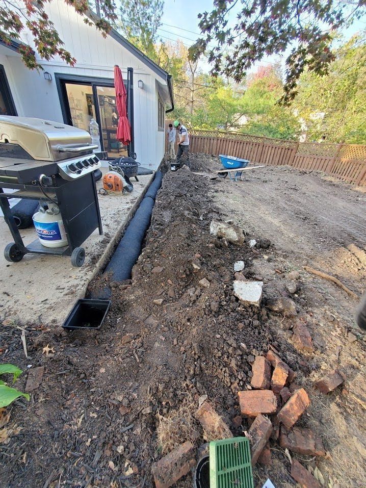 Backyard excavation beside house with exposed trench, dirt pile, bricks, and a grill on the patio