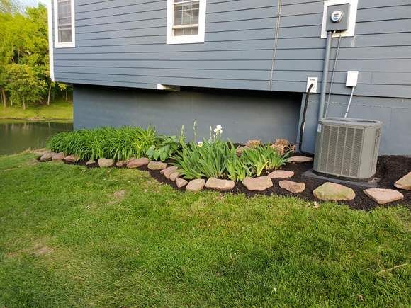 House foundation with landscaped flower bed, rocks, green plants, and an air conditioner unit beside a lawn.