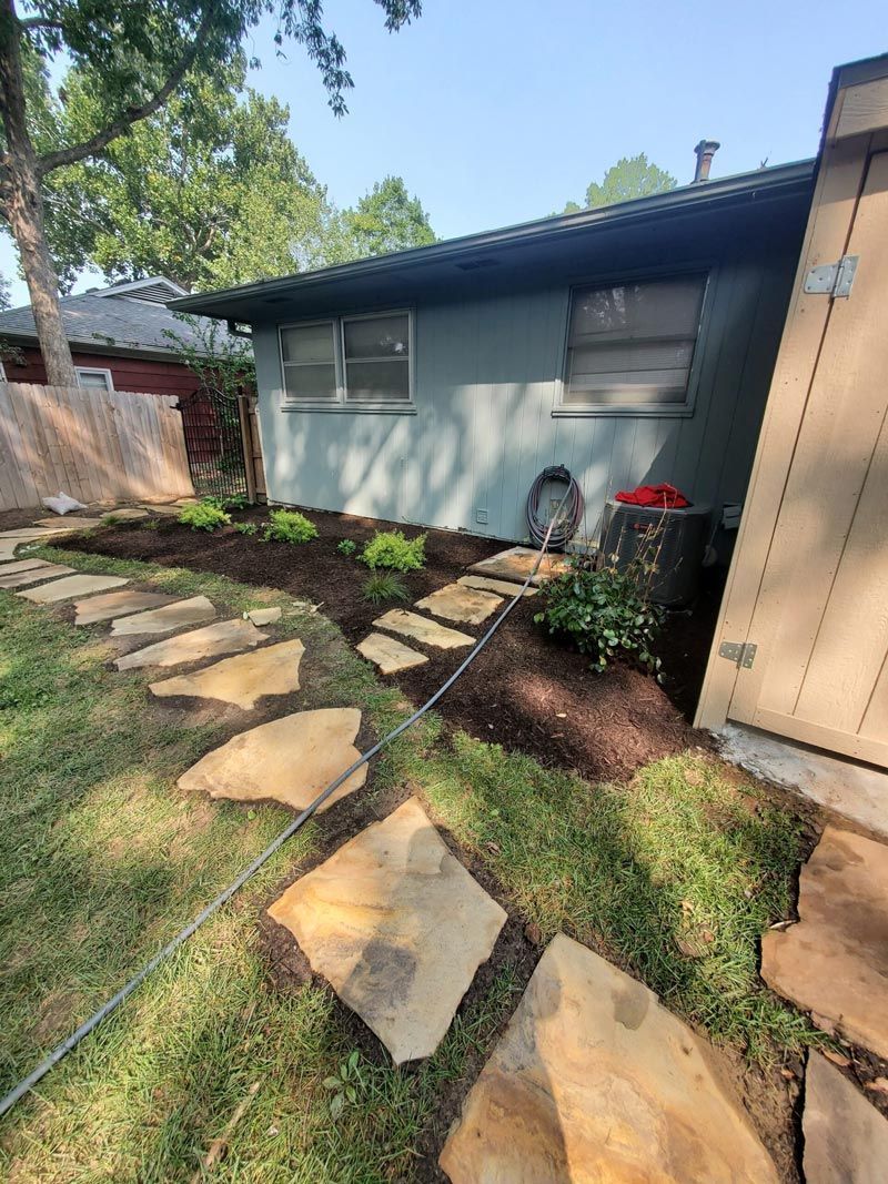 Stone path through a grassy yard beside a light blue house with a tan shed and trees.