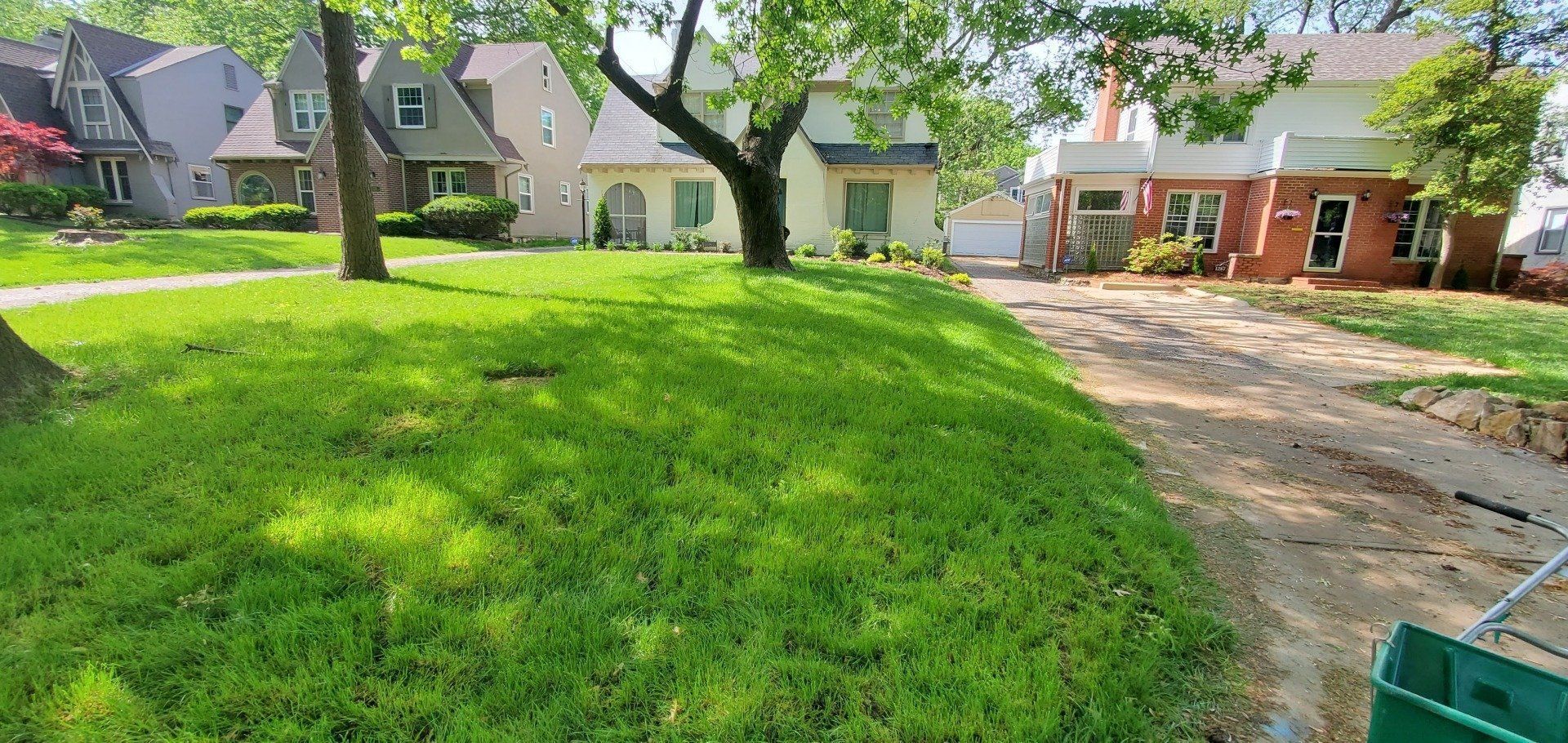 Suburban front yard with bright green lawn, trees, and houses beside a gravel driveway