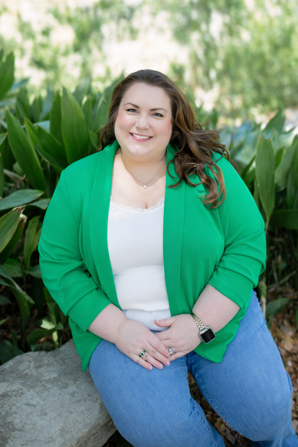 Woman in a green blazer and blue jeans sitting on a rock outdoors, smiling.