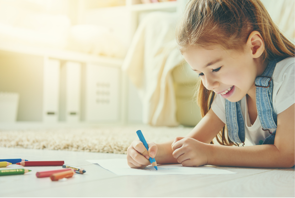 Girl drawing with a blue pencil on paper, smiling, on a rug in a bright room.
