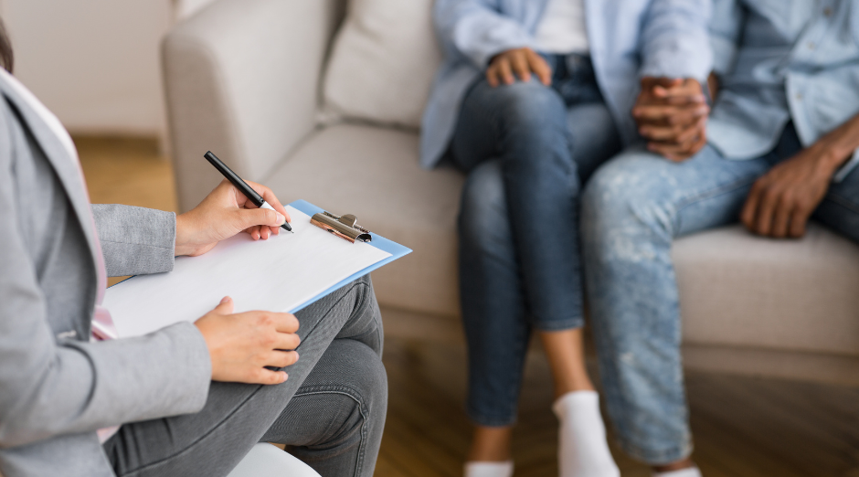 A picture of a couple holding hands on a sofa with a woman taking notes across from them