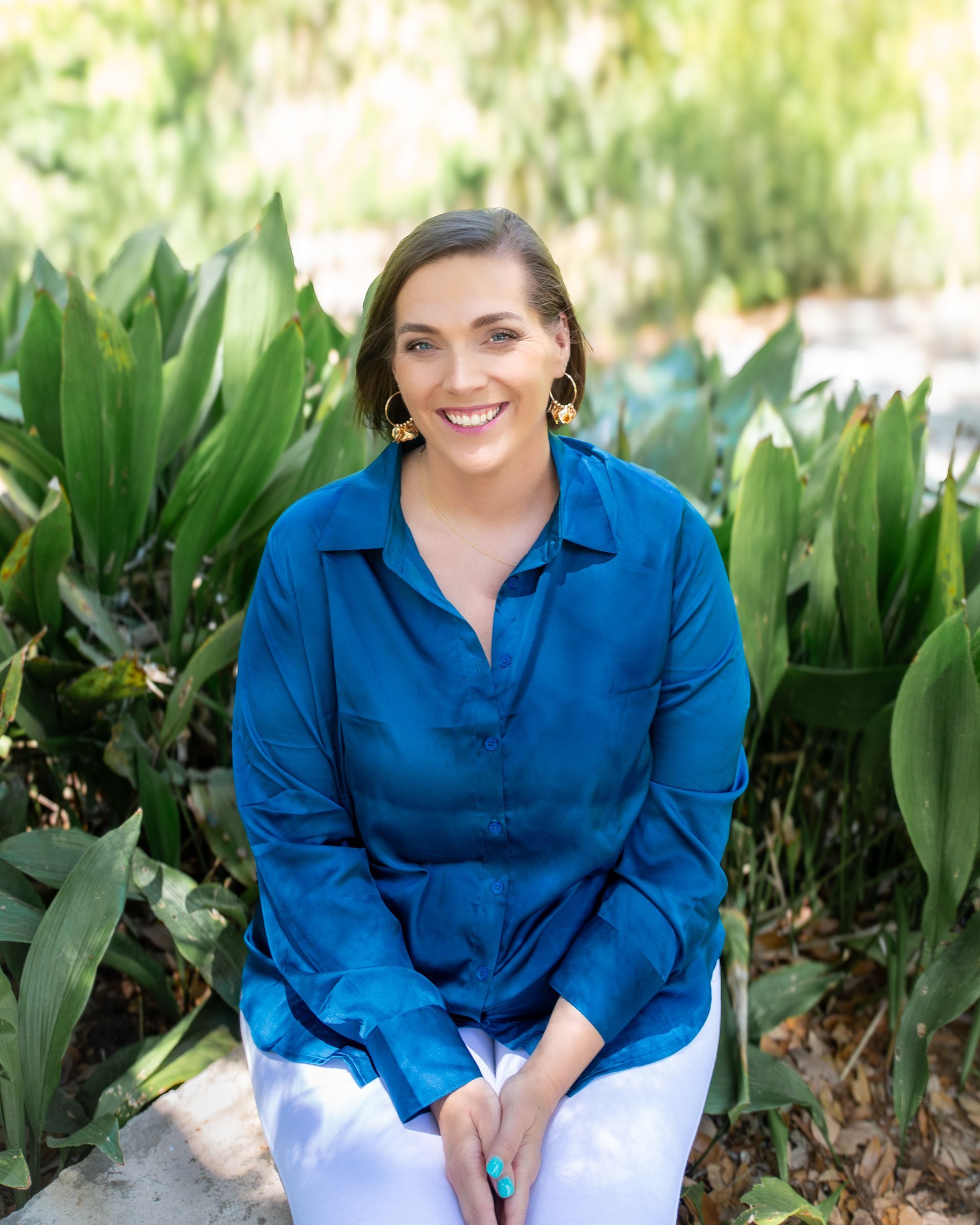 Smiling woman in a blue blouse sits outdoors among green plants.