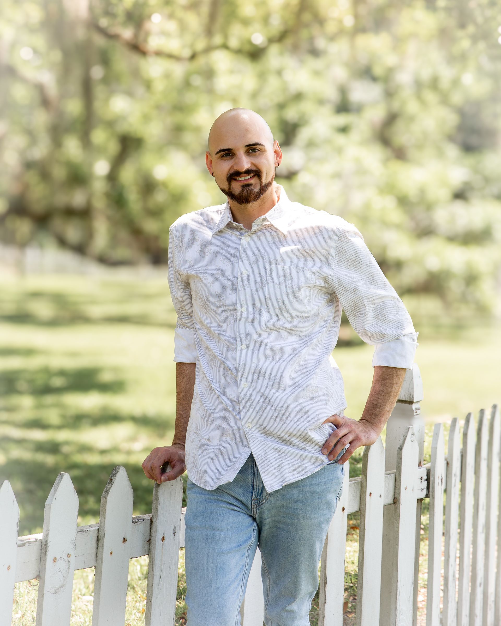 Man leaning on a white picket fence in a sunny park, wearing a white shirt and light jeans.