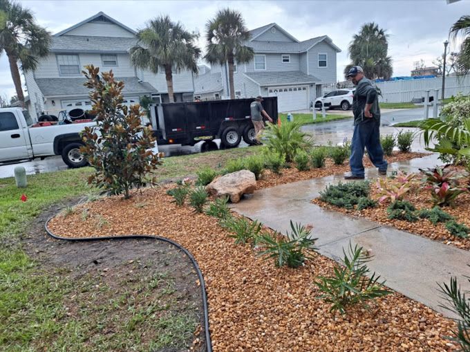 Landscapers working on a front yard with fresh mulch and plants, near a trailer and houses.