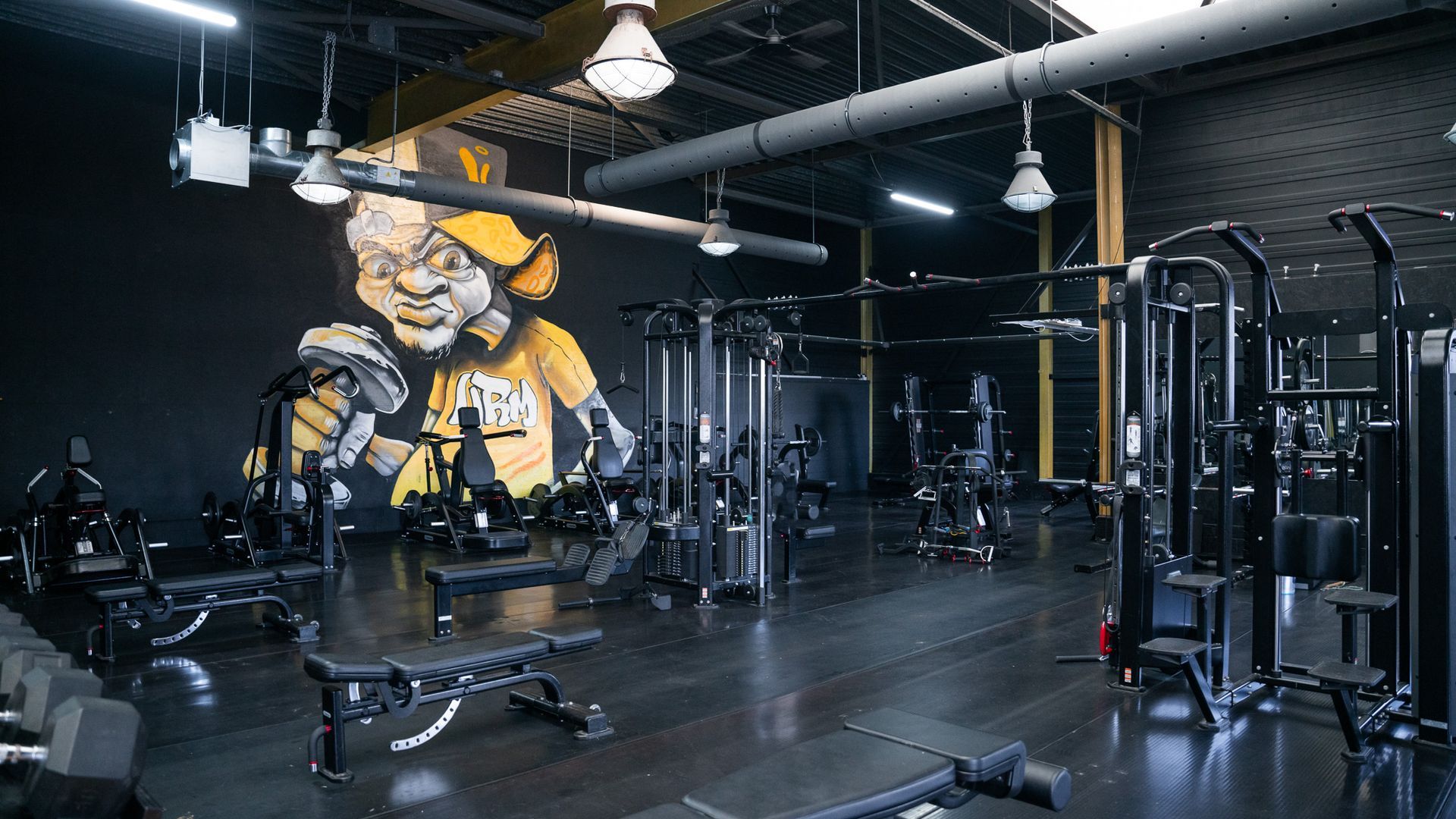 Interieur van een sportschool met donkere muren, diverse fitnessapparaten en een grote geel-zwarte muurschildering.