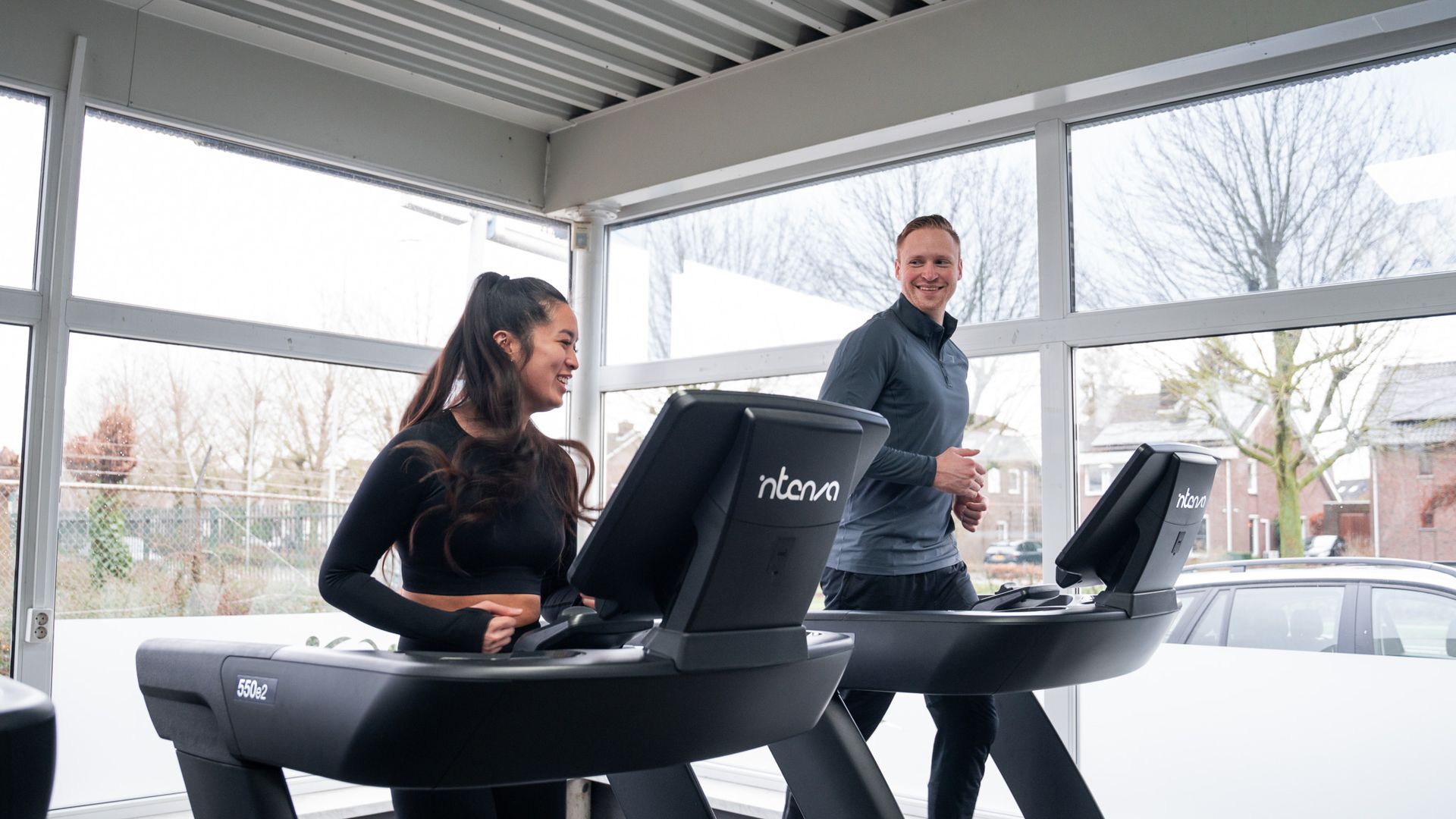 Twee mensen op loopbanden in een sportschool met uitzicht op de besneeuwde buitenlucht. Ze zijn glimlachend aan het sporten.