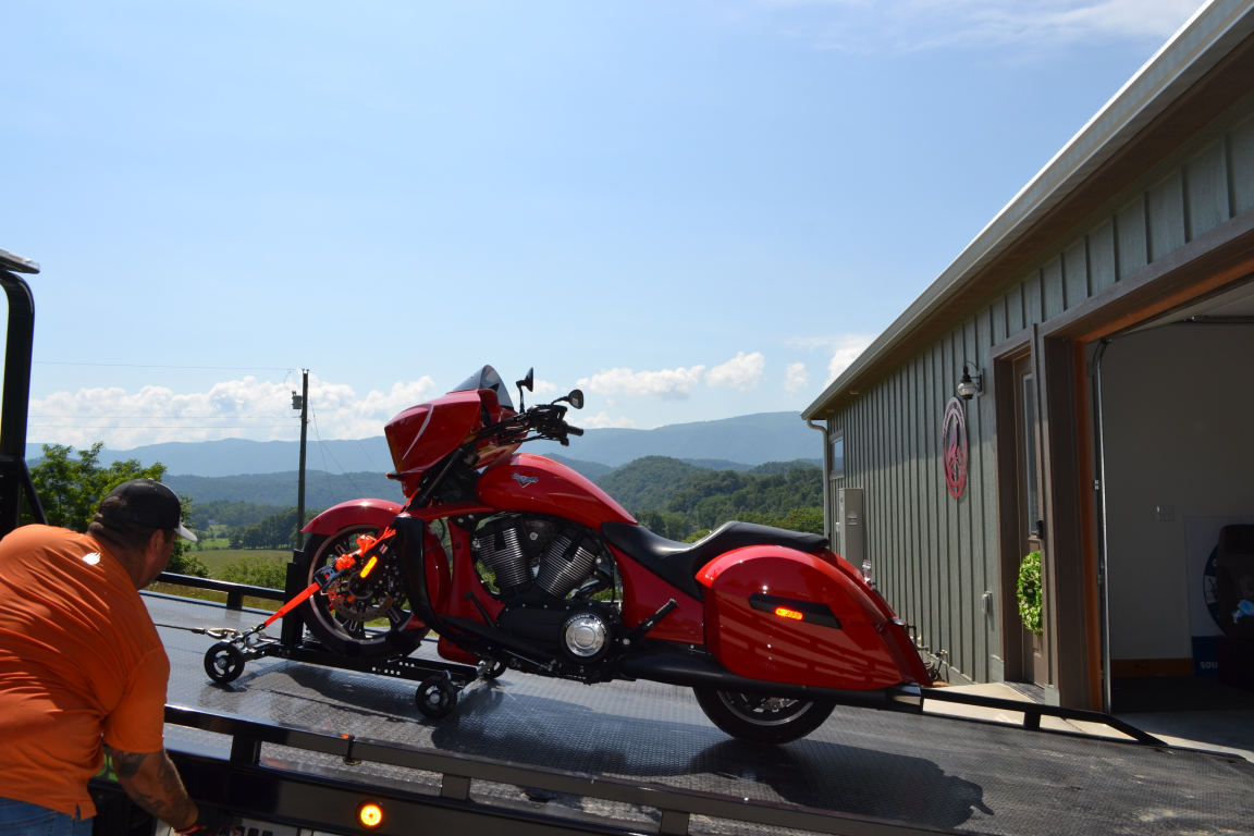 Red custom motorcycle on a trailer beside a building, with another bike and mountains in the background.