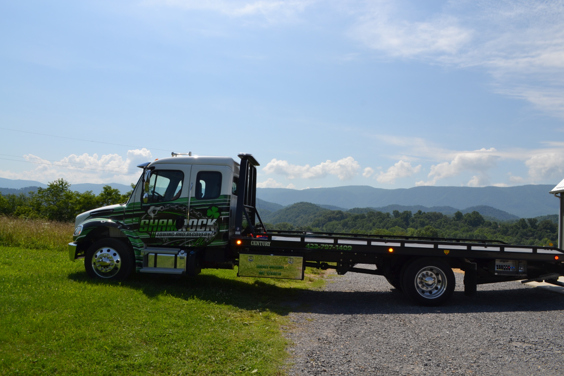 Flatbed tow truck parked on grass beside a road with mountains and blue sky in the background