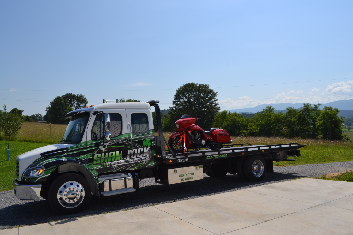 Flatbed tow truck with green graphics carrying a red motorcycle on a sunny roadside.