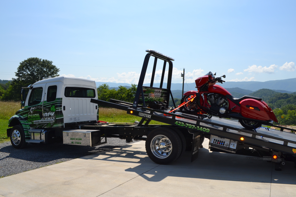 White flatbed tow truck carrying a red motorcycle on a sunny road with mountains in the background