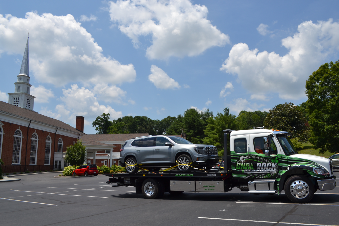 Tow truck carrying a silver SUV beside a red-brick building with a church steeple under a cloudy sky