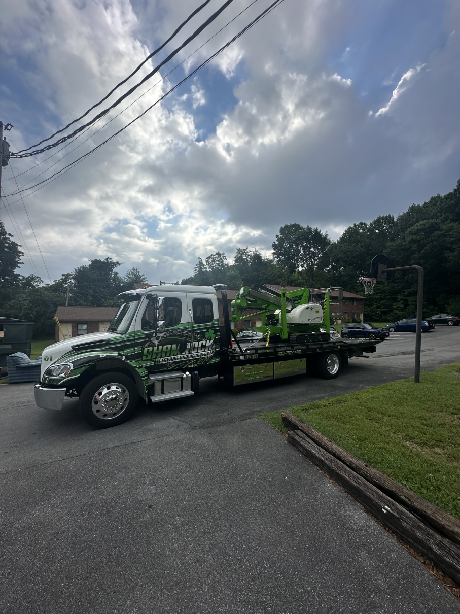 Tow truck carrying a damaged green car parked on a residential street under cloudy skies