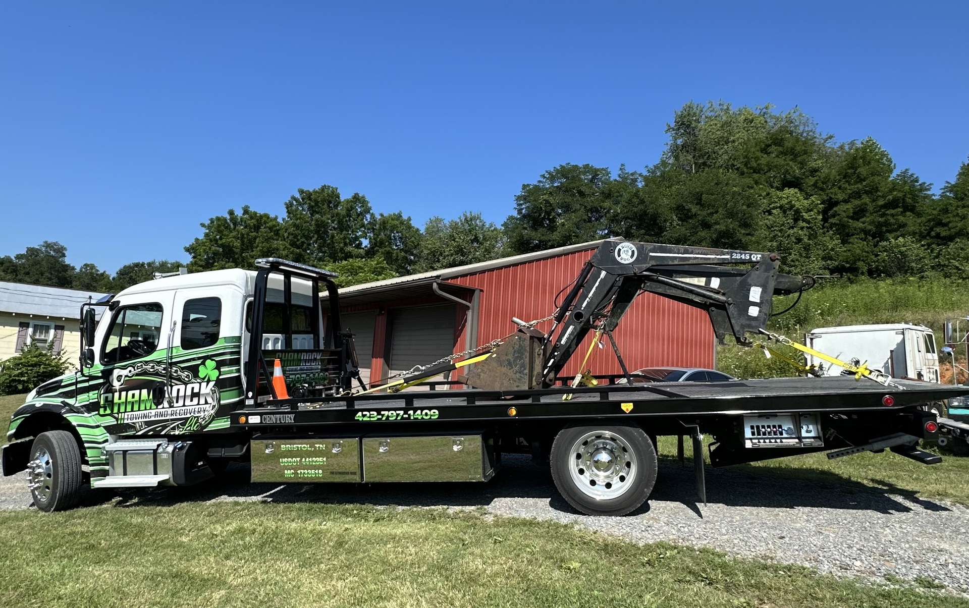 Tow truck with flatbed and boom parked outdoors beside a red barn under a blue sky