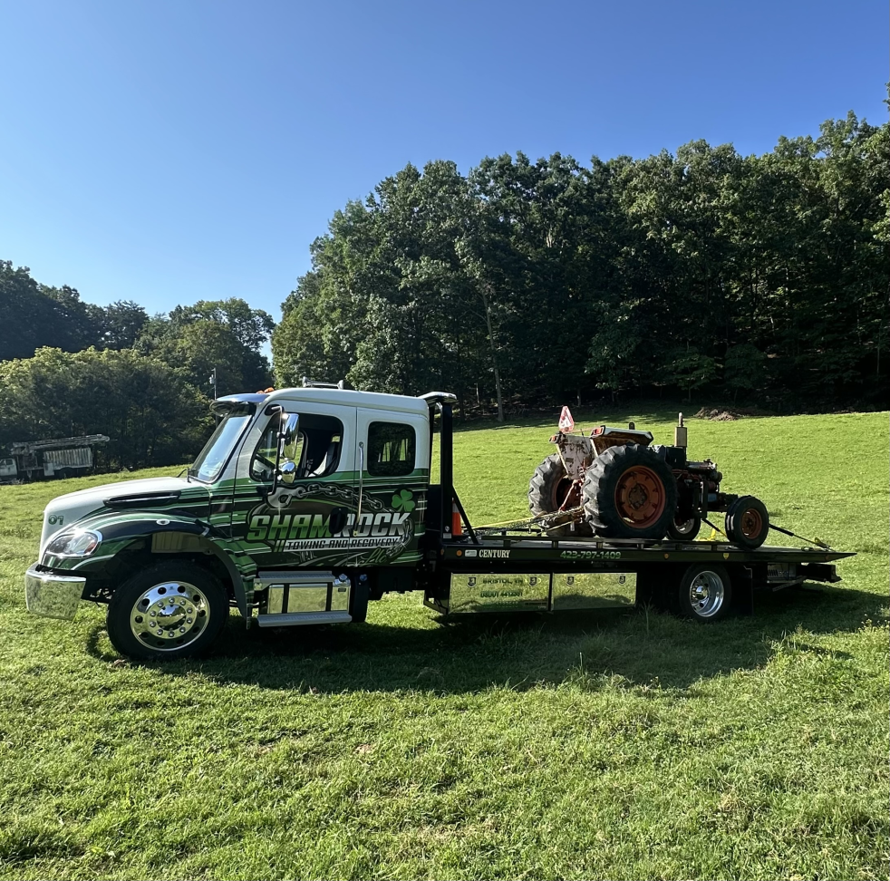 Flatbed tow truck carrying a rusty tractor in a grassy field under a clear blue sky