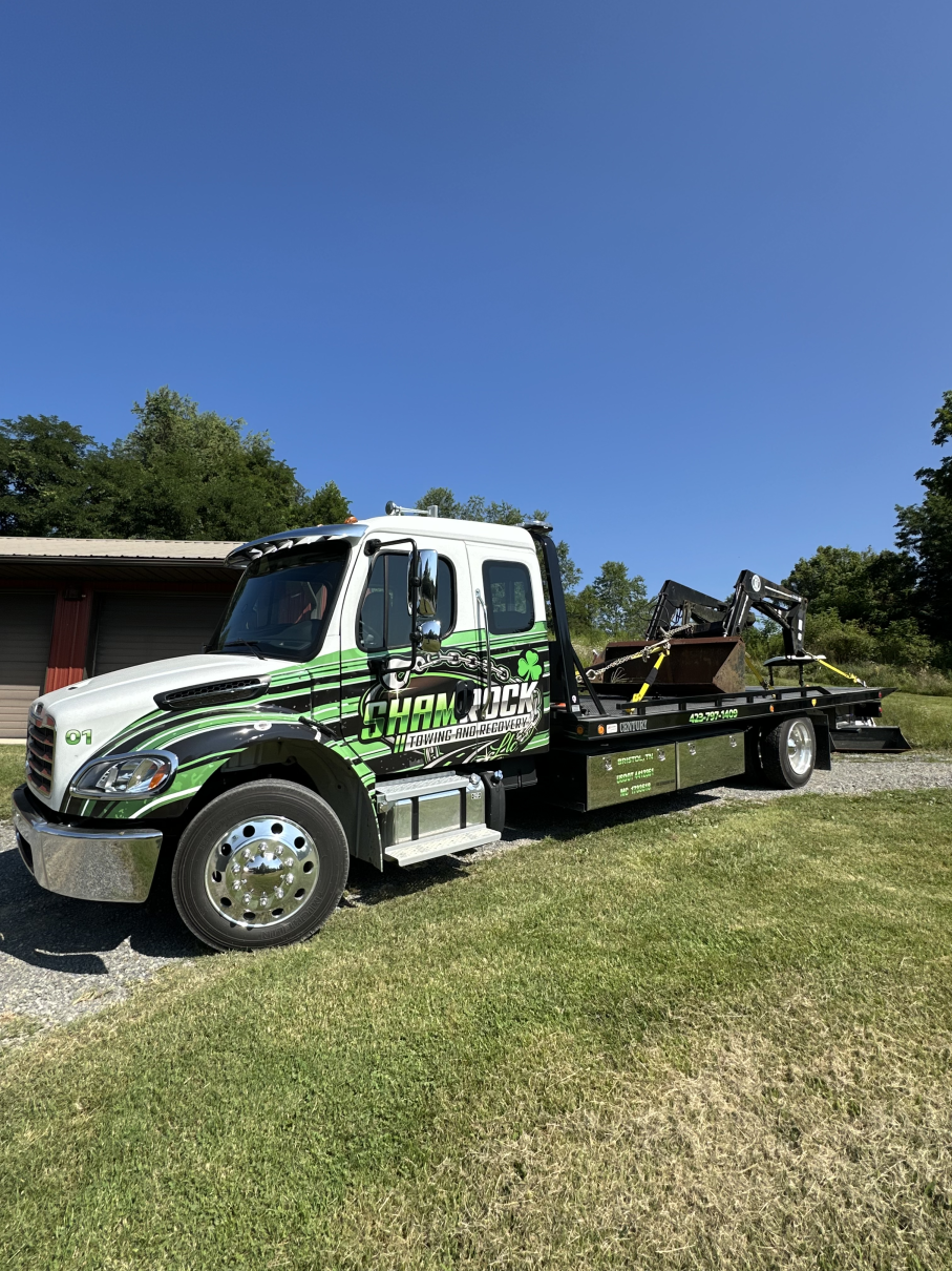Green and white tow truck parked on grass beside a house under a clear blue sky