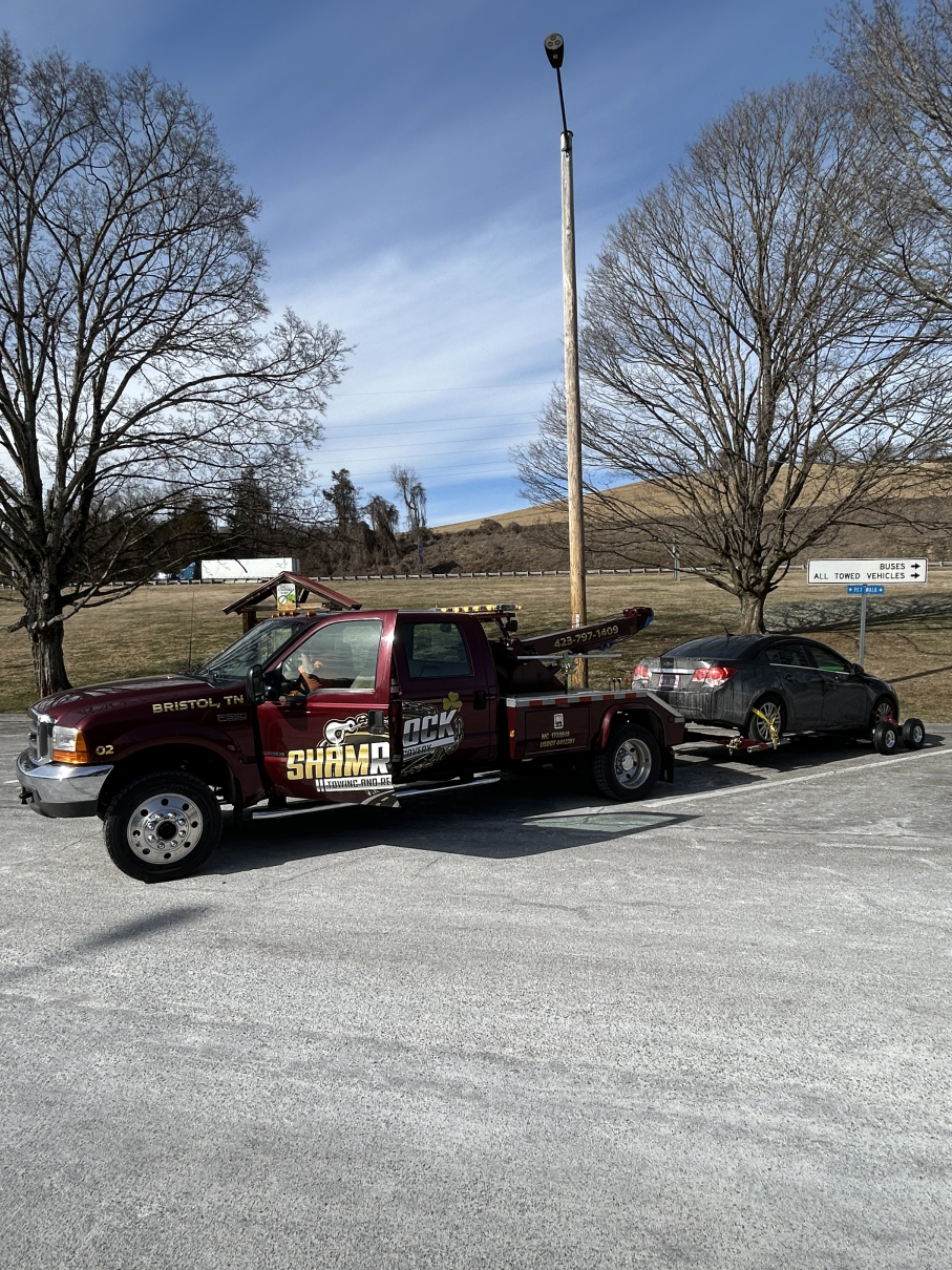 Tow truck hauling a black sedan in a gravel lot with bare trees and a lamppost.