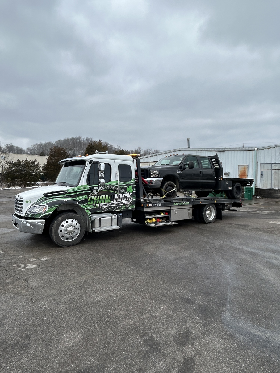 Camouflage tow truck carrying a black pickup in a gravel lot under a cloudy sky