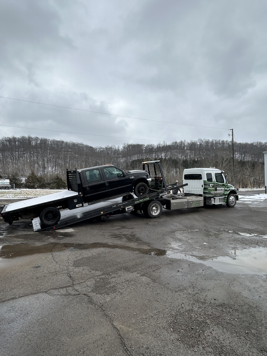 Tow truck hauling a dark pickup on a flatbed in a wet parking lot under cloudy skies
