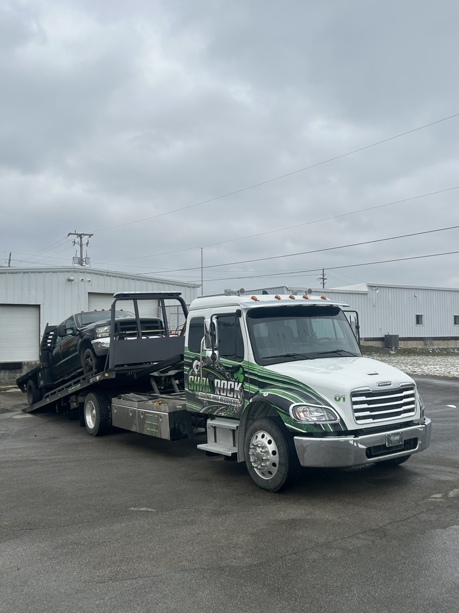 Flatbed tow truck carrying a damaged black SUV in an industrial lot under cloudy skies