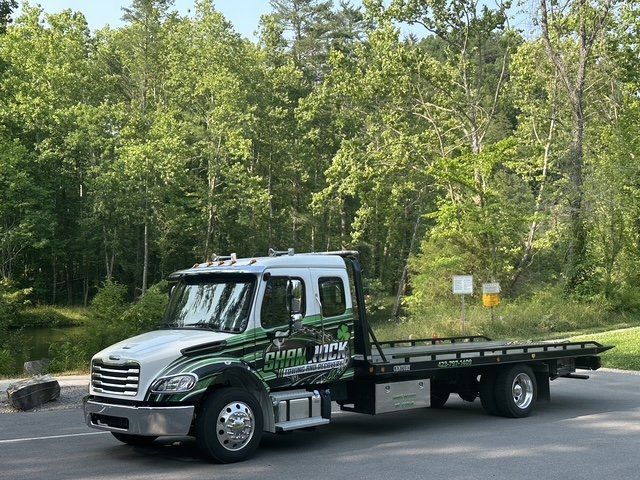 Green flatbed tow truck parked on a road beside a wooded area
