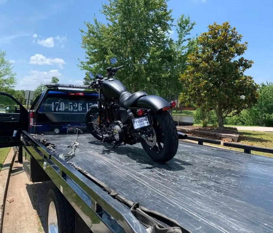 Black motorcycle on a flatbed tow truck in a sunny outdoor lot with trees.