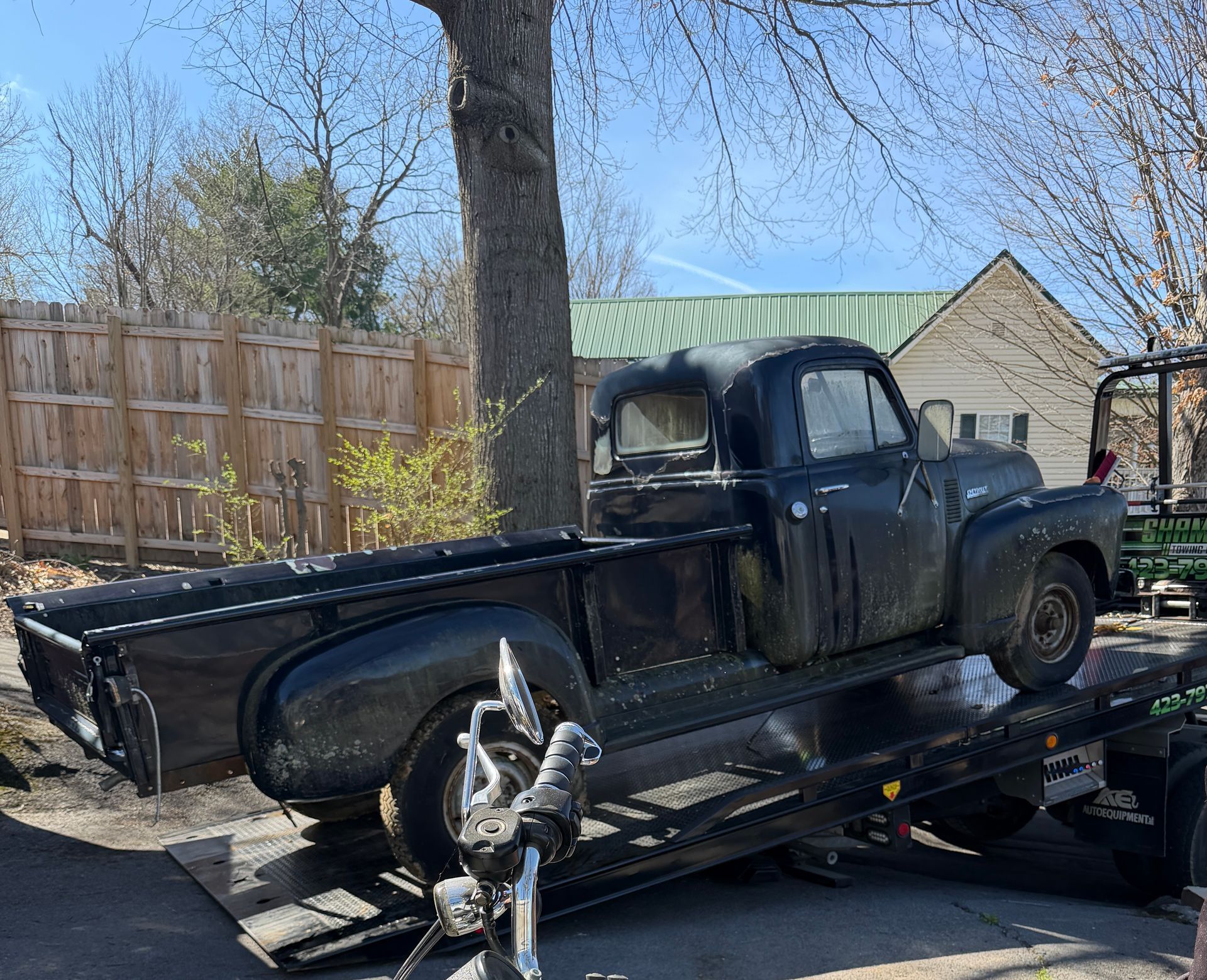 Vintage black pickup truck on a flatbed trailer in a sunny backyard with a fence and tree.
