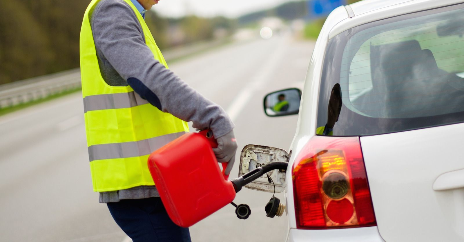 Roadside worker in a reflective vest fueling a white car from a red gas can