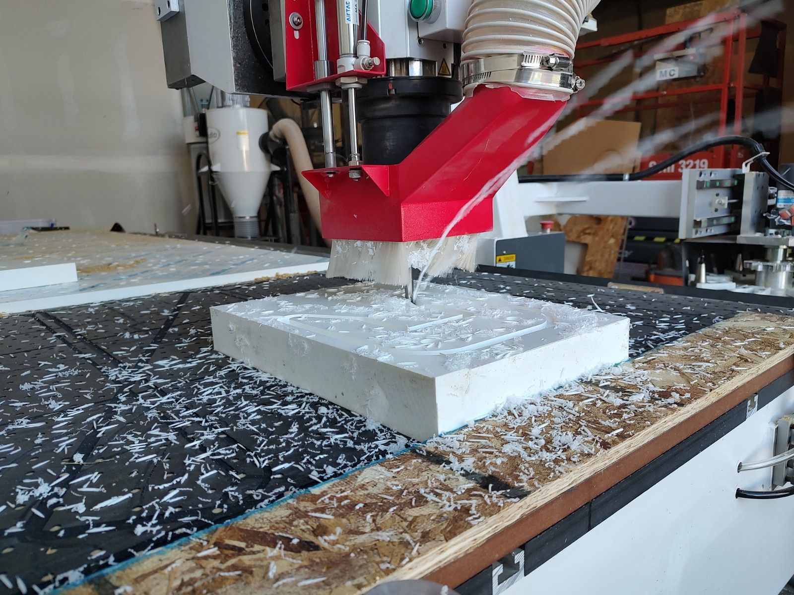 CNC router cutting a white foam block on a workbench with red dust extraction hose nearby