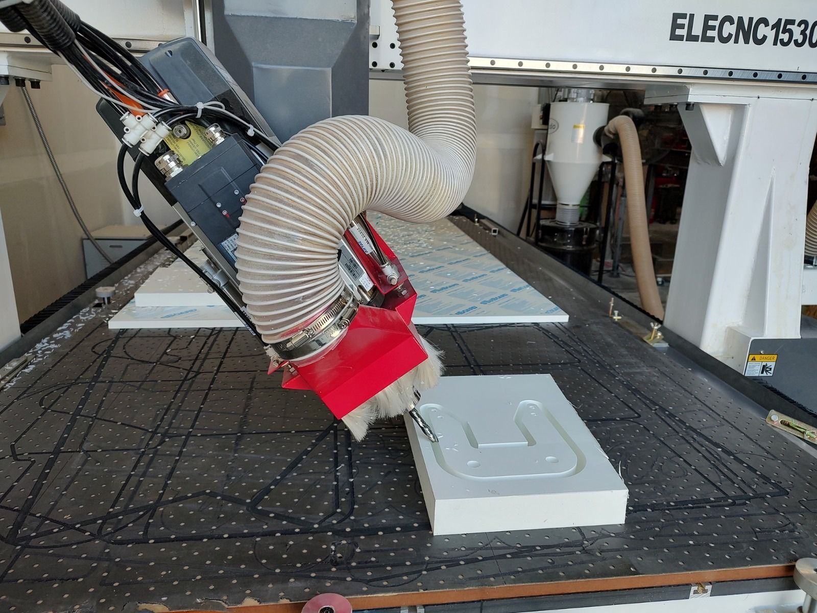 CNC router cutting a light gray panel with a red spindle and dust hose in a workshop