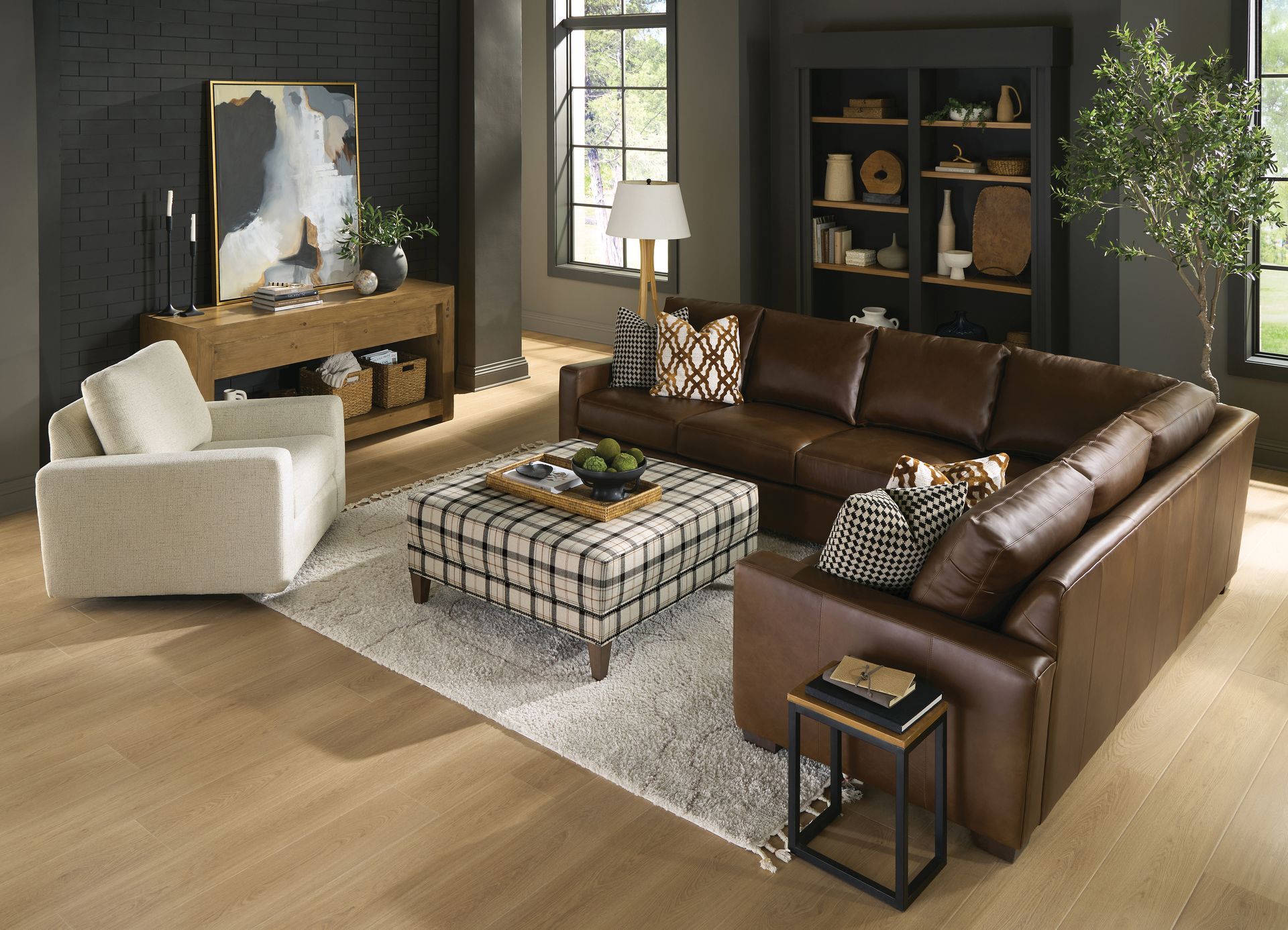 Living room with brown leather sectional, white armchair, plaid ottoman, and decor.