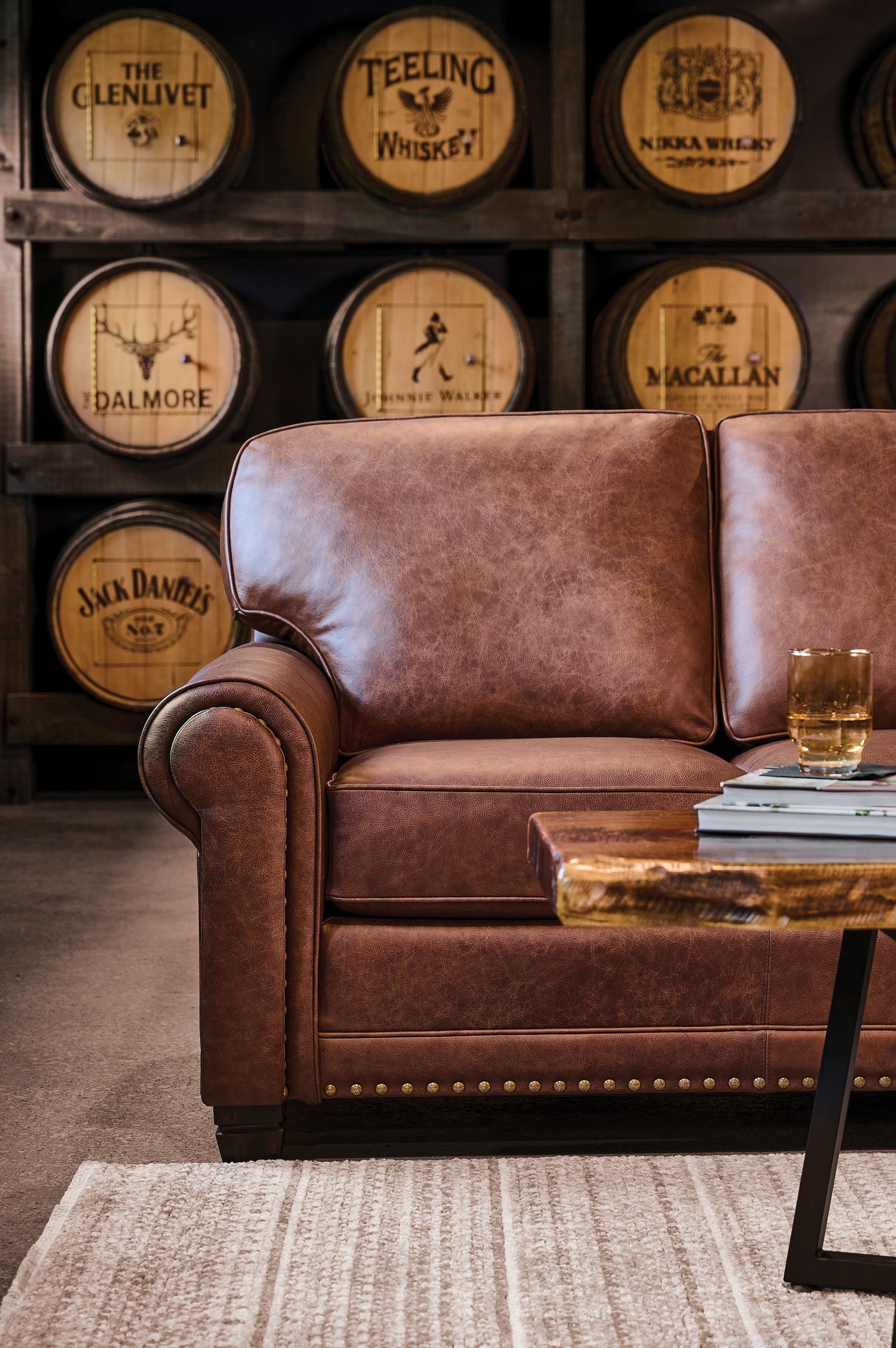 Brown leather couch in front of whiskey barrel wall, with a small wood and metal table and a drink.