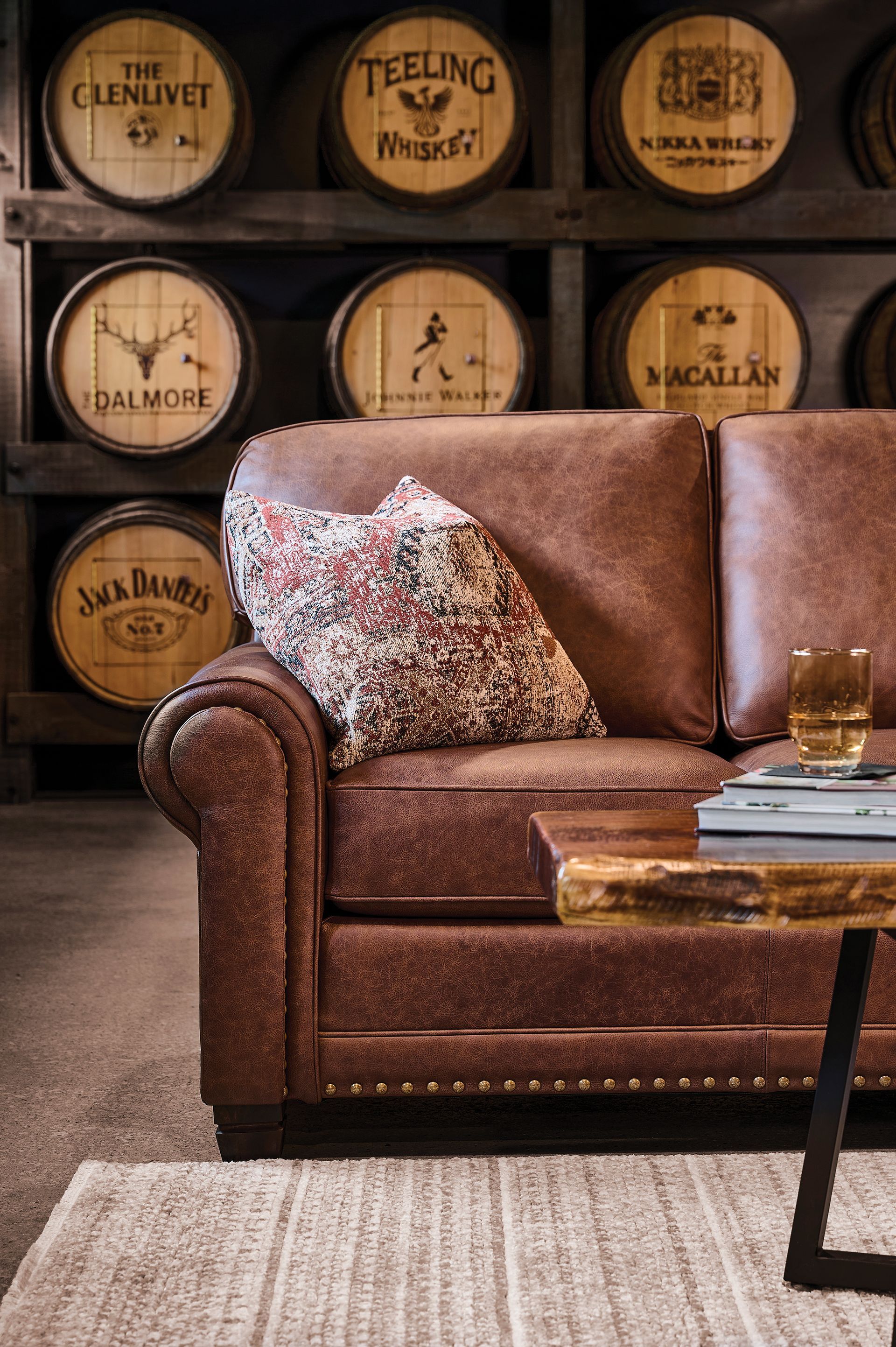 Brown leather couch with patterned pillow, wooden side table, and whiskey barrels in the background.