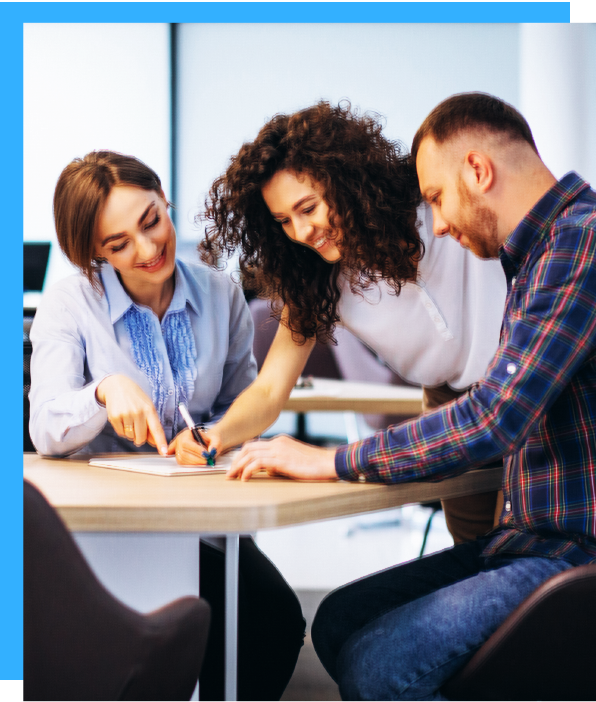 Three people collaborate around a table, one points to a document while smiling, office setting.