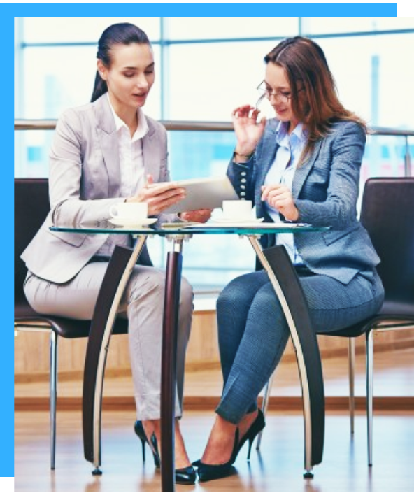 Two businesswomen in suits sit at a glass table, reviewing a tablet and papers in a modern office.