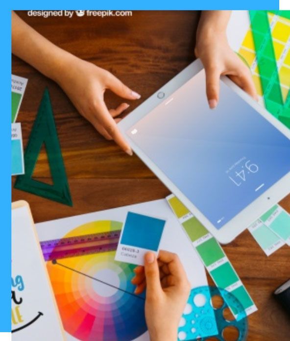 Hands working on design elements: tablet, color swatches, ruler, color wheel, and green triangle on a wooden table.