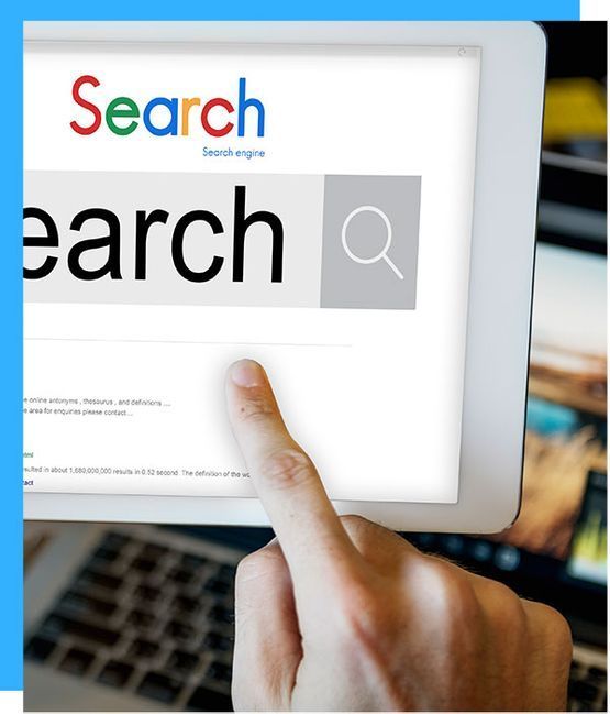 Finger pointing at a search bar on a tablet screen, search icon. Laptop visible in background.