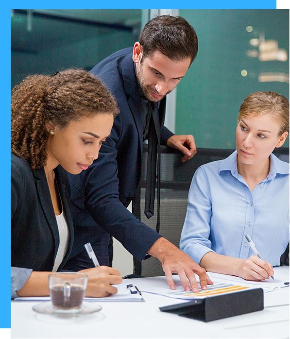 Three business colleagues reviewing documents at a table. A man points at a paper, and two women write.