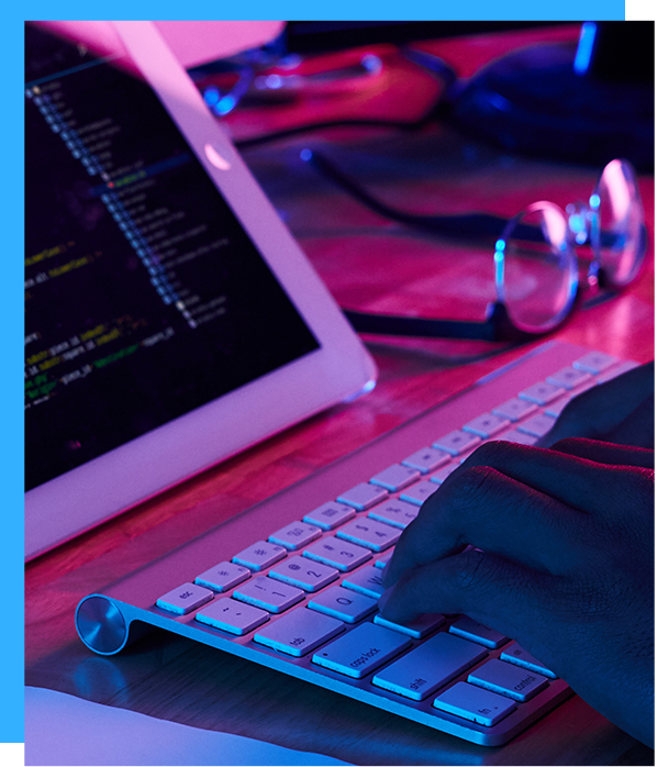 Person typing on a keyboard, with a tablet displaying code. Glasses rest on the desk, lit by blue and pink light.