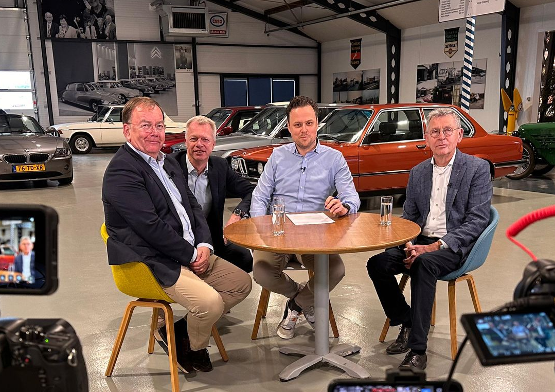Four people sitting around a round table in a car showroom with vintage vehicles in the background.