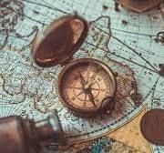 An antique brass compass rests open on top of an old, detailed world map, accompanied by a telescope lens.